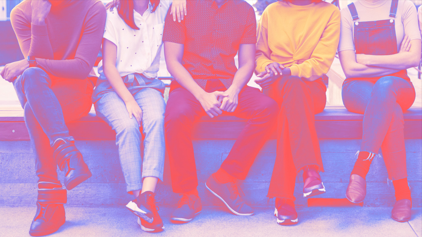 Six young adult friends sitting in a row on a bench in a city street smiling to camera, full length
